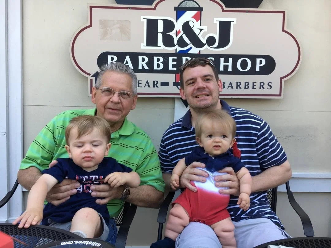 Two men, each holding a baby, sitting in front of the R&J Barber Shop sign